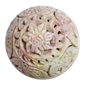 Puede incluir: Una esfera de piedra tallada con intrincados diseños florales. La esfera es una mezcla de tonos rosa claro, beige y gris. El diseño presenta tallas detalladas de flores y hojas, creando un efecto calado.