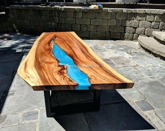 May include: A wooden table with a live edge design. The tabletop features a river of bright blue epoxy resin flowing through the centre, contrasting with the natural wood grain. The table is supported by a black metal base.