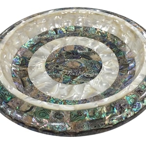 Op de afbeelding: Een decoratieve, ronde kom met een mozaïekontwerp. Het heeft concentrische cirkels van witte, iriserende en grijsgroene abalone schelpstukken. De rand en de basis zijn donkergrijs, wat een opvallend contrast creëert.