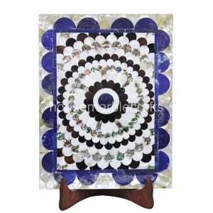 May include: A rectangular decorative piece with a floral design. The piece features a central circular pattern in black, white, and iridescent materials, surrounded by a scalloped border in blue and white. It is displayed on a wooden stand.