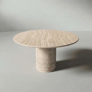 May include: A round table featuring a beige travertine stone top with natural veining and a cylindrical base. The base is constructed from the same beige stone and displays a vertical fluted texture along its central section.