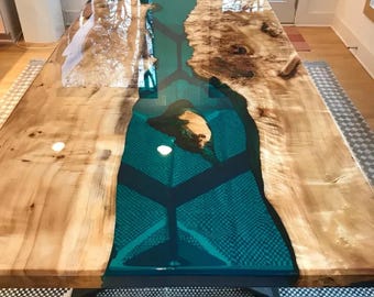Puede incluir: Una mesa de madera rectangular grande con un río de resina epoxi turquesa que atraviesa el centro. La madera tiene un color marrón claro natural con nudos y vetas de color marrón oscuro. La mesa se apoya sobre una base de metal negro.