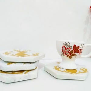 Puede incluir: Posavasos de mármol blanco con diseños dorados de ciervos. Una taza de té blanca con detalles florales rojos descansa sobre un posavasos a juego. Flores rojas decorativas en un jarrón blanco.