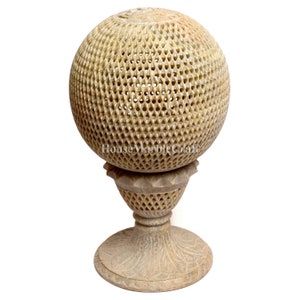 Puede incluir: Una esfera de piedra beige tallada con un diseño de celosía, que descansa sobre una base de pedestal a juego. La esfera tiene un patrón calado detallado. La base presenta intrincadas tallas florales, creando una pieza decorativa.