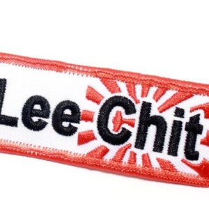 Könnte beinhalten: Weißer Schlüsselanhänger mit rotem Rand und einem schwarzen gestickten Design, das "Hoo Lee Chit" mit einem roten und weißen Sonnenstrahl-Design sagt.