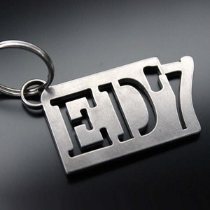 Puede incluir: Un llavero de metal plateado con un diseño recortado que dice "ED'7".