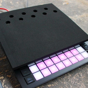 Pode incluir: Um pad de música eletrônica preto com uma capa de espuma preta. O pad tem 16 botões iluminados e alguns botões com rótulos de texto, incluindo "Função", "Bateria" e "Presets".