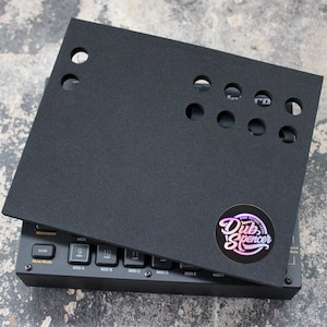 Può includere: Cuscino in schiuma nera con ritagli per manopole e pulsanti. Il cuscino è progettato per adattarsi sopra un controller di produzione musicale. Il cuscino ha un adesivo con il testo "Dub Spencer" in un cerchio.