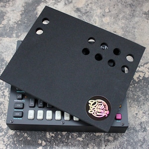 Può includere: Cuscino in schiuma nera con ritagli per manopole e pulsanti, progettato per proteggere un sintetizzatore musicale. Il cuscino è posto sopra un sintetizzatore nero con un adesivo bianco che dice "Dub Spencer".