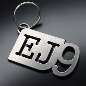 Può includere: Un portachiavi in metallo argentato con le lettere "EJ9" tagliate. Il portachiavi ha un anello in metallo argentato.