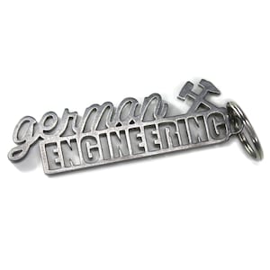Könnte beinhalten: Ein silberner Schlüsselanhänger aus Metall mit den Worten "german engineering" und einem stilisierten Schraubenschlüssel- und Hammerdesign.