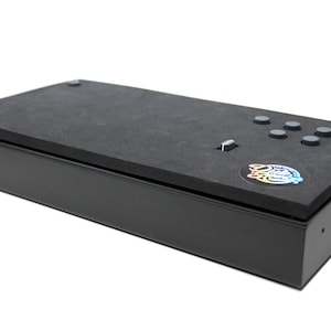 Può includere: Una scatola rettangolare nera con un top in schiuma nera. Il top in schiuma ha un adesivo circolare con un design colorato e diversi pulsanti neri.