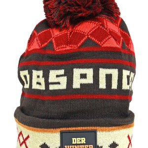 Könnte beinhalten: Braune Strickmütze mit rotem und weißem geometrischem Muster, einem roten Bommel oben und einem schwarzen Patch mit dem Text "DER WINTER GERÄT" auf der Vorderseite.