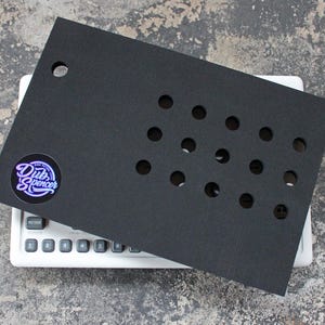 Può includere: Un pannello nero rettangolare con molteplici fori circolari, che copre parzialmente un dispositivo elettronico bianco con pulsanti. Il pannello presenta un adesivo circolare con il testo "Dub Spencer".