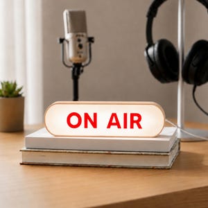 May include: An oval-shaped light box with a light wood frame and a white front panel featuring the text ON AIR in bold red letters. The sign is resting on top of two stacked white books, positioned in front of a microphone and black headphones.