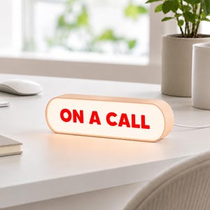 Könnte beinhalten: Ein rechteckiges, pillenförmiges Schreibtischschild mit abgerundeten Kanten und einem hellen Rahmen. Die Vorderseite ist beleuchtet und zeigt den Text ON A CALL in fetten, roten Großbuchstaben. Ein dünnes weißes Kabel führt an der rechten Seite heraus.