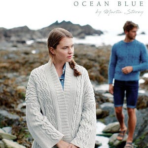 May include: A light gray cable-knit cardigan with a shawl collar and long sleeves. The text at the top reads ROWAN OCEAN BLUE by Martin Storey. A blue cable-knit sweater and dark blue shorts are visible in the background on a rocky shore.