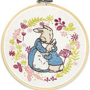 Puede incluir: Arte de aro bordado con dos conejos abrazándose, rodeados por una corona floral. La corona presenta acentos rosas, amarillos y burdeos. El bordado está en un aro de madera.