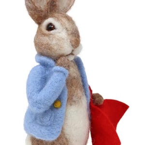 Puede incluir: Un conejo de fieltro marrón y blanco que lleva una chaqueta azul y una capa roja. El conejo tiene orejas largas y una mancha blanca en el pecho.