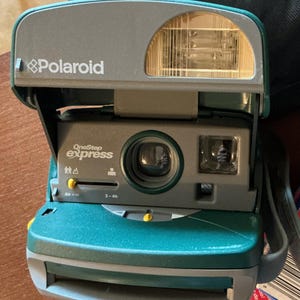 May include: A teal and gray Polaroid OneStep Express instant camera featuring a built-in flash, a central lens, and a viewfinder. The front panel displays the text Polaroid and OneStep Express with focus distance settings ranging from 2 feet to infinity.