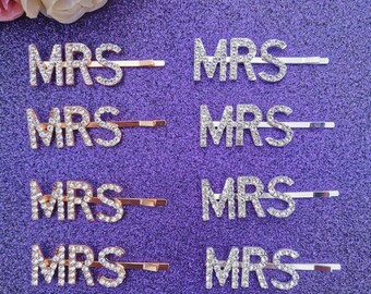 Könnte beinhalten: Sechs goldene Haarklammern mit dem Wort "MRS" in Strasssteinen. Die Klammern befinden sich auf einem glitzernden lila Hintergrund.