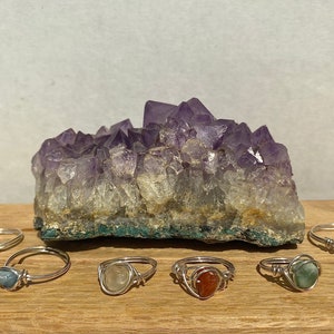 May include: Seven silver rings with different colored gemstones. The rings are arranged on a wooden surface in front of a large amethyst crystal.