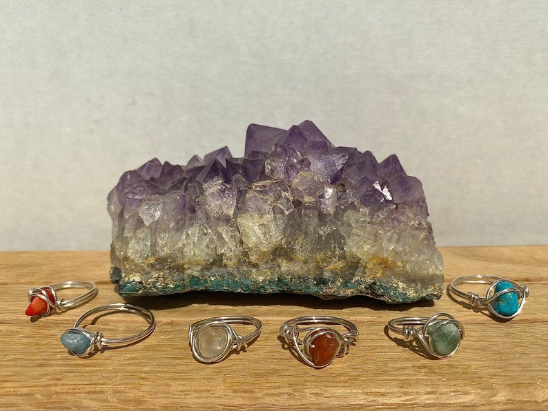 Peut inclure: Sept bagues en argent avec des pierres pr&eacute;cieuses de diff&eacute;rentes couleurs. Les bagues sont dispos&eacute;es sur une surface en bois devant un grand cristal d'am&eacute;thyste.