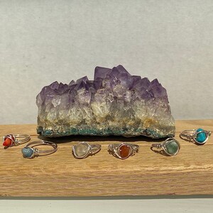 Peut inclure: Sept bagues en argent avec des pierres pr&eacute;cieuses de diff&eacute;rentes couleurs. Les bagues sont dispos&eacute;es sur une surface en bois devant un grand cristal d'am&eacute;thyste.