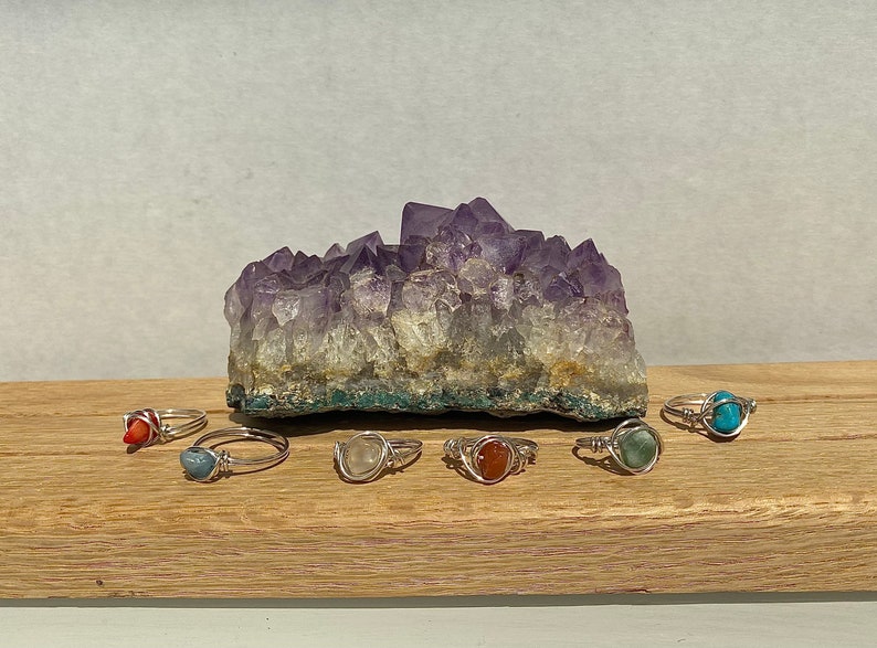 Peut inclure: Sept bagues en argent avec des pierres pr&eacute;cieuses de diff&eacute;rentes couleurs. Les bagues sont dispos&eacute;es sur une surface en bois devant un grand cristal d'am&eacute;thyste.