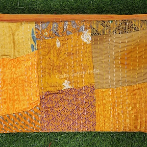 Könnte beinhalten: Eine Patchwork-Decke aus verschiedenen Stoffen in Orange- und Brauntönen. Die Decke ist handgenäht mit weißem Garn und zeigt eine Vielzahl von Mustern, darunter florale, geometrische und Paisley-Muster. Die Decke liegt auf einer grünen Grasfläche. "Crafts Central India" ist auf die Decke gedruckt.