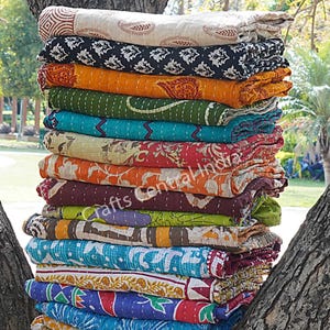 May include: A stack of colorful, patterned quilts rests between tree trunks. The quilts display a variety of designs, including paisley, geometric shapes, and floral patterns. The color palette includes shades of blue, green, orange, and red.