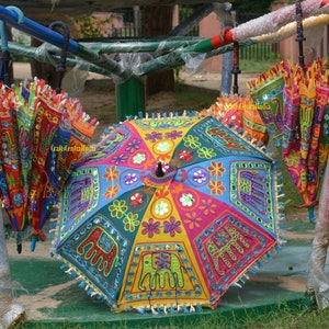 Puede incluir: Tres paraguas coloridos con intrincados bordados y detalles de flecos. Los paraguas están hechos de tela y tienen una variedad de patrones, incluyendo elefantes, flores y diseños geométricos. Los paraguas están colgados de un marco de metal.