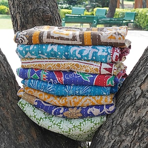 Könnte beinhalten: Ein Stapel von acht bunten Kantha-Steppdecken mit verschiedenen floralen und geometrischen Mustern. Die Decken sind aus recycelten Saris hergestellt und handgenäht. Sie sind gefaltet und auf einem Baumstamm gestapelt.