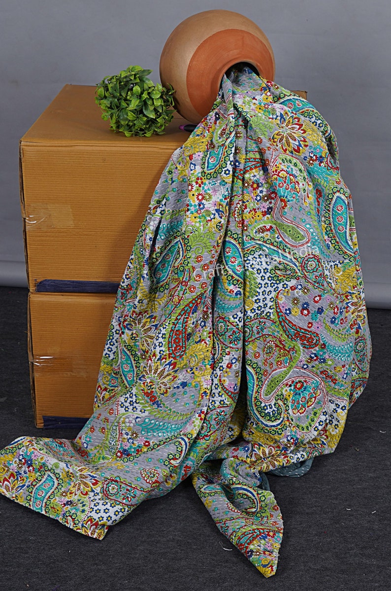 May include: A grey and white kantha quilt with a paisley pattern in shades of green, blue, red and yellow. The quilt is draped over a cardboard box and a terracotta pot.