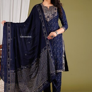 May include: A navy blue three-piece outfit with gold embroidery. The outfit includes a long-sleeved top, trousers, and a matching dupatta. The top has a round neckline and a decorative pattern on the front. The trousers are straight-legged and the dupatta is a sheer fabric with a border.