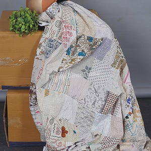 Könnte beinhalten: Ein Patchwork-Quilt mit einer Vielzahl von Stoffquadraten mit Mustern in neutralen Tönen, darunter Weiß, Beige und Grau. Der Quilt ist über einer Holzkiste drapiert, mit einem Terrakotta-Topf und einer kleinen grünen Pflanze im Hintergrund. Das Design umfasst florale und geometrische Muster.