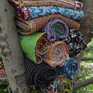 May include: A stack of colorful kantha quilts rolled up and tied with string, displayed against a tree trunk. The quilts are made of various fabrics and feature intricate embroidery and patchwork designs.