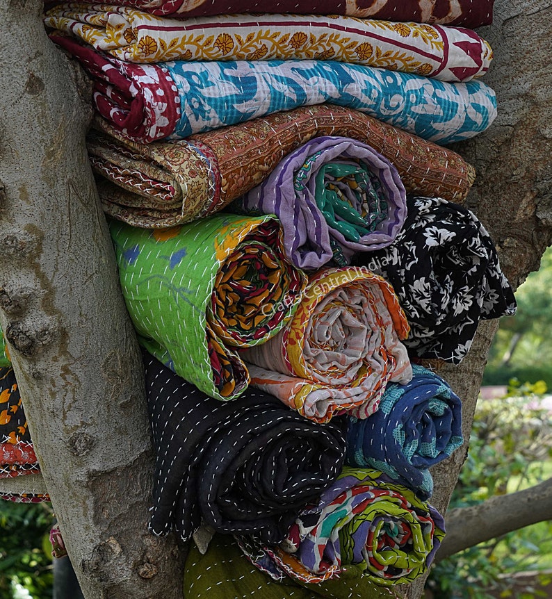 May include: A stack of colorful kantha quilts rolled up and tied with string, displayed against a tree trunk. The quilts are made of various fabrics and feature intricate embroidery and patchwork designs.