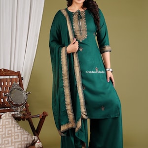 May include: A green kurta with gold embroidery and a matching dupatta. The kurta has a round neckline and three-quarter sleeves. The dupatta has a gold border. The outfit is worn by a woman standing in front of a green background.