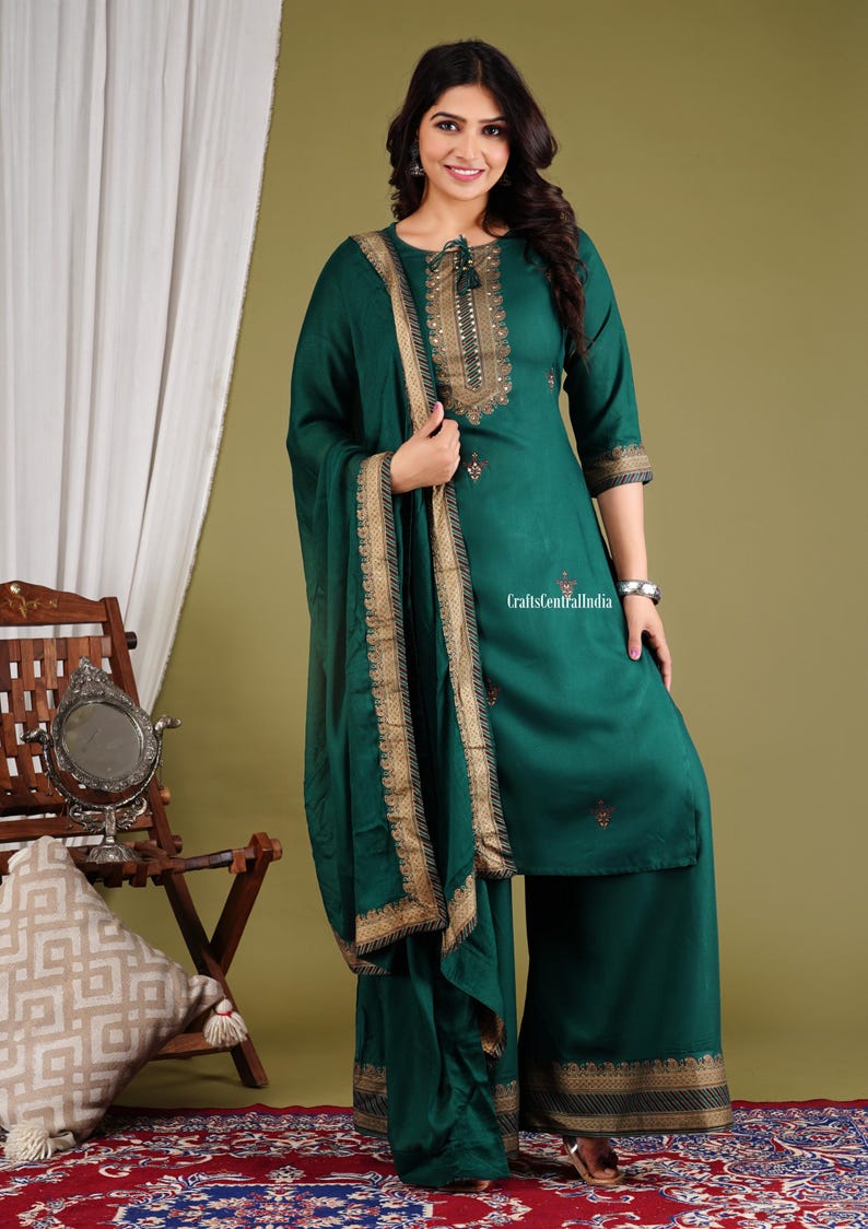 May include: A green kurta with gold embroidery and a matching dupatta. The kurta has a round neckline and three-quarter sleeves. The dupatta has a gold border. The outfit is worn by a woman standing in front of a green background.