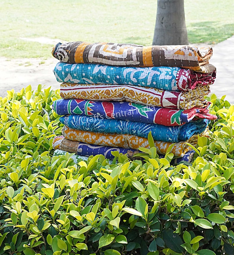 May include: A stack of six colorful kantha quilts, made from recycled saris, with intricate patterns and stitching. The quilts are folded and stacked on top of each other, creating a visually appealing display.