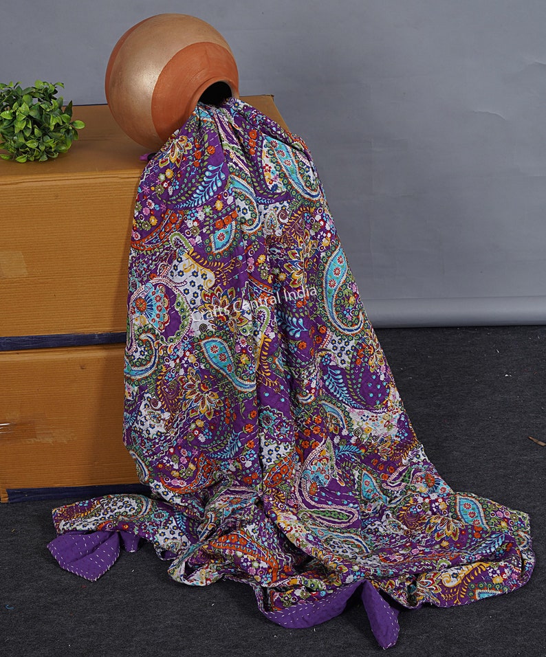 May include: A purple kantha quilt with a paisley pattern and colourful floral accents. The quilt is draped over a brown box and a terracotta pot.