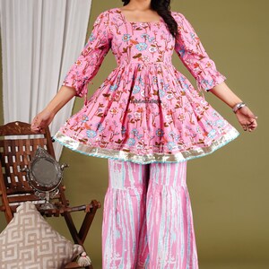 Op de afbeelding: Een roze en witte, met bloemen bedrukte katoenen kurta met een uitlopende vorm en 3/4 mouwen. De kurta heeft een geplooide taille en een zilveren rand. Het model draagt de kurta met bijpassende roze en witte palazzo broek.