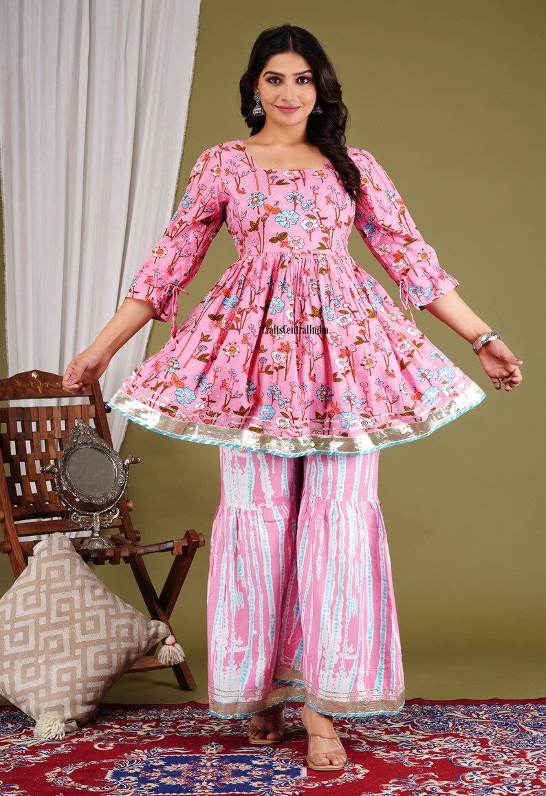 Op de afbeelding: Een roze en witte, met bloemen bedrukte katoenen kurta met een uitlopende vorm en 3/4 mouwen. De kurta heeft een geplooide taille en een zilveren rand. Het model draagt de kurta met bijpassende roze en witte palazzo broek.