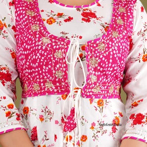 May include: White floral print top with a pink embroidered jacket. The jacket has a tie closure and a white tassel. The top has a round neckline and three-quarter sleeves.