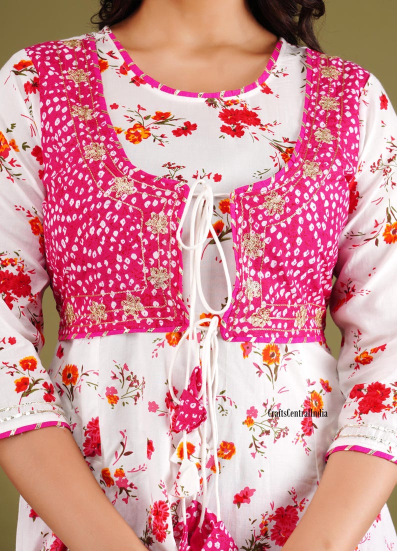 May include: White floral print top with a pink embroidered jacket. The jacket has a tie closure and a white tassel. The top has a round neckline and three-quarter sleeves.