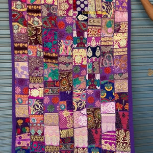 Könnte beinhalten: Ein großer Wandteppich mit Patchwork-Muster. Der Wandteppich besteht aus verschiedenen Stoffen in den Farben Lila, Rosa, Gold und Blau. Der Stoff ist mit Stickereien und Pailletten verziert. Der Wandteppich hat einen lilafarbenen Rand.