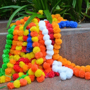 Könnte beinhalten: Eine bunte Girlande aus künstlichen Blumen, hergestellt aus Pompons in den Farben Grün, Gelb, Orange, Rot, Weiß und Blau. Die Girlande besteht aus mehreren Blumensträngen, die ein dickes und festliches Aussehen erzeugen.
