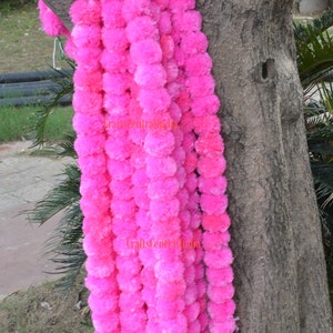 Könnte beinhalten: Eine lange Girlande aus rosa Stoffblumen, auch bekannt als Ringelblumengirlanden, die an einem Baumstamm hängt. Die Girlande besteht aus vielen kleinen, flauschigen, rosa Pompons.