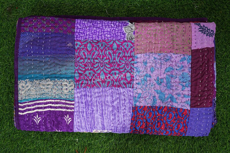 May include: A patchwork quilt made from repurposed saris in shades of purple, pink, blue, and red. The quilt is hand-stitched and has a variety of floral and geometric patterns.